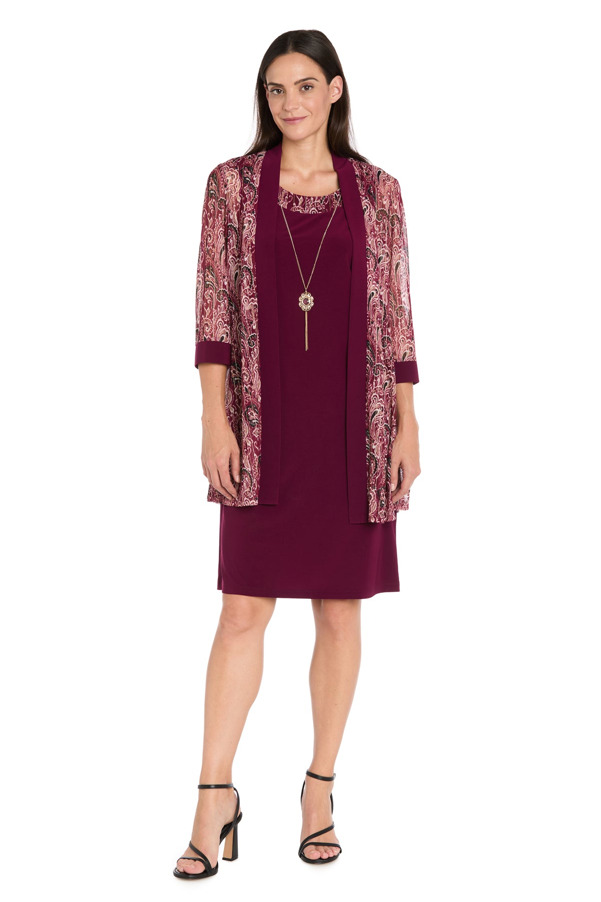 A woman in the Petite Short Wine Party Dress: R&M Richards 3029P, styled with a paisley-patterned jacket, black heeled sandals, and a long necklace, stands against a white background.